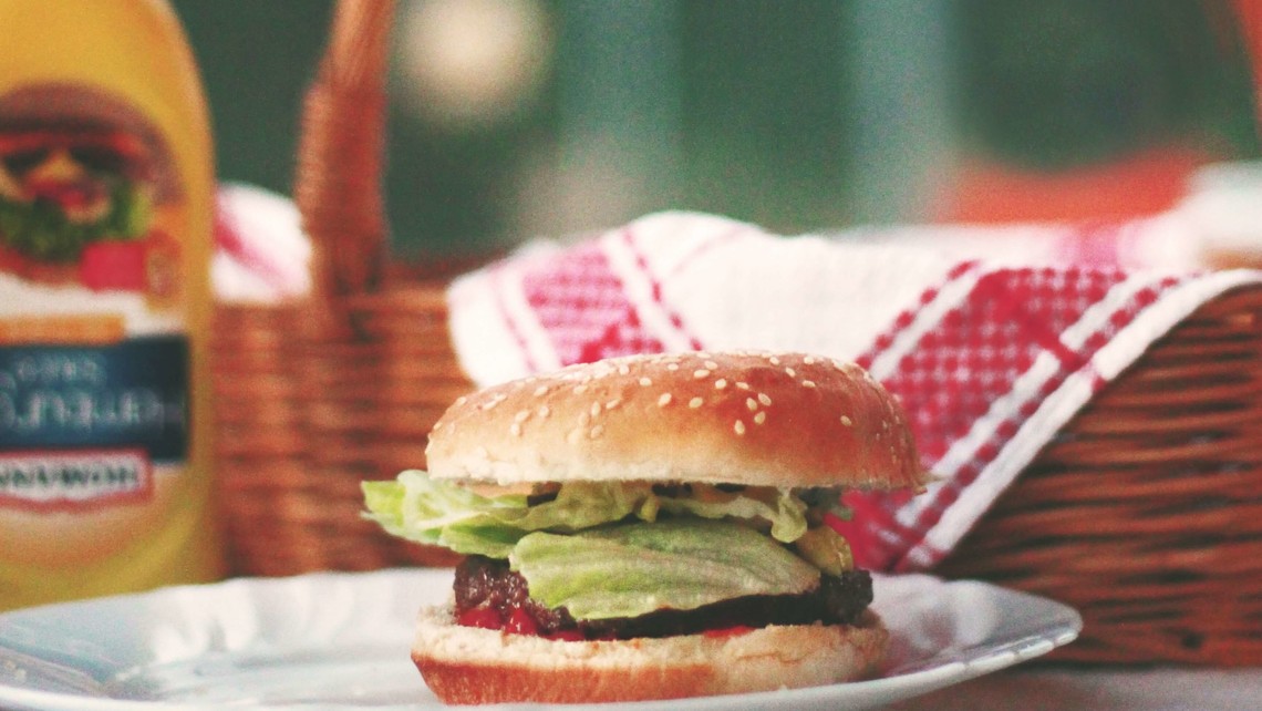 hamburger sits on a plate with mustard and straw basket in the background